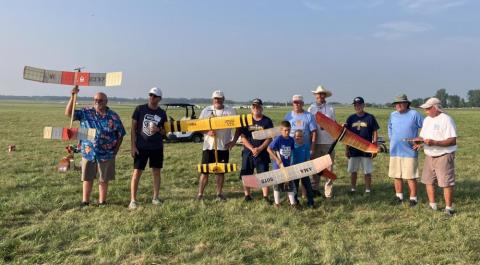 People holding model planes on a grassy field, smiling, under a clear blue sky.