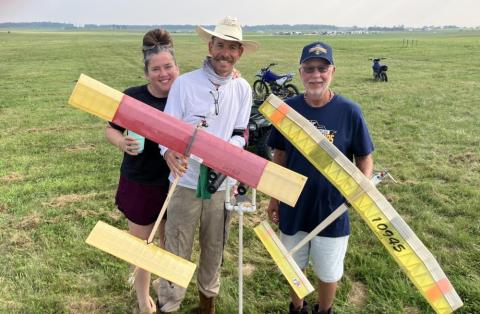 Three people smiling in a grassy field holding model airplanes.