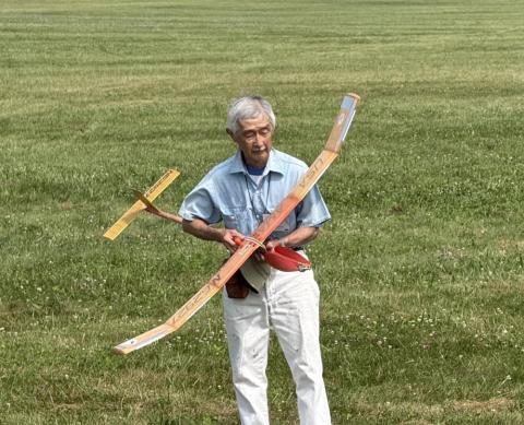 Elderly man holding a model glider on a grassy field.