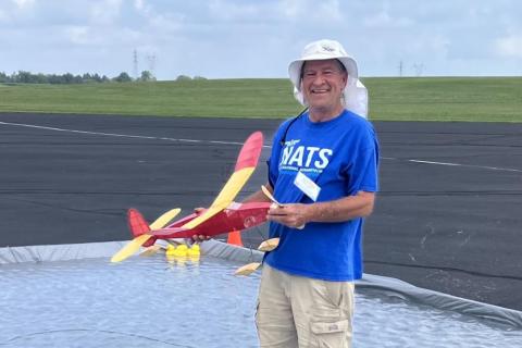Man in blue shirt holding model airplane by a pond on an airfield.