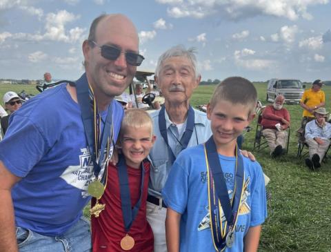 Group of four people smiling outdoors, wearing medals, with a cloudy sky background.
