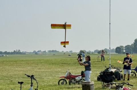 Flying model airplane with orange and yellow wings on a grassy field.