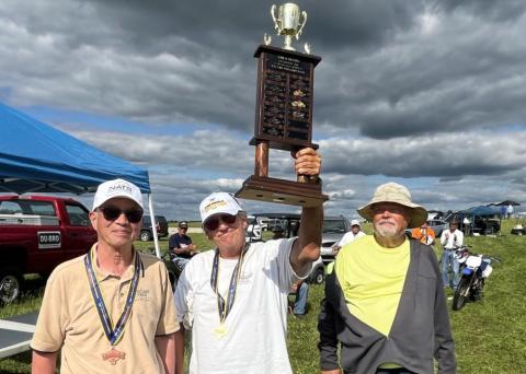 Three men outdoors, one holding a tall trophy under a cloudy sky.