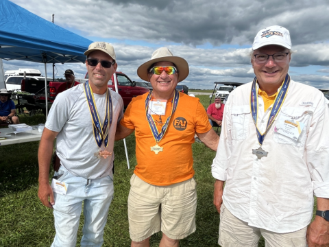 Three men outdoors wearing medals, smiling under a cloudy sky.