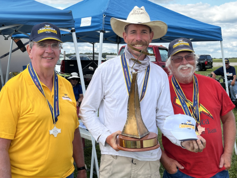 Three men with medals; one holding a trophy, smiling under blue tents.