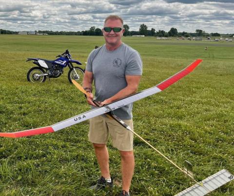 Man holding a model glider on a grassy field, motorcycle in the background.