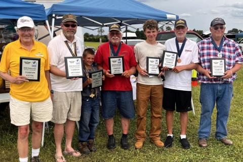 Seven people holding plaques under blue canopies at an outdoor event.