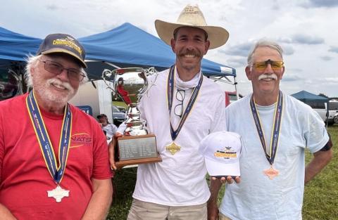 Three men in casual attire, wearing medals, smile outdoors. One holds a trophy.