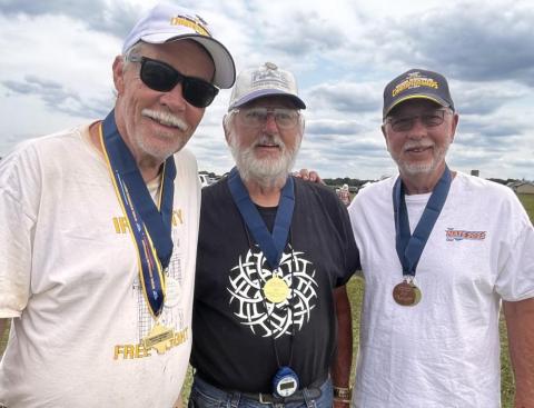 Three men outdoors wearing medals, smiling under a cloudy sky.