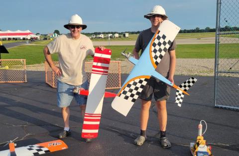 Two people smiling, holding colorful model airplanes at an airfield.