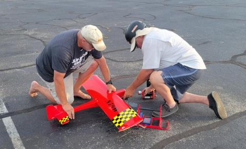 Two men kneel to assemble a red model plane on pavement.