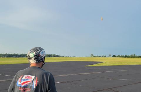 Man with helmet watches small model aircraft fly over an airfield.