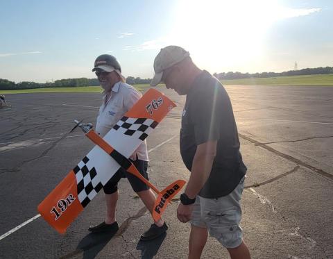 Two men walking on a runway, holding a model airplane with orange and checkered wings.