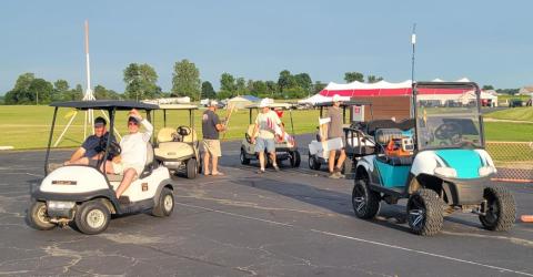 Golf carts on a paved road, people standing nearby, sunny day.