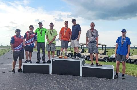 Seven people standing on a podium outdoors with golf carts in the background.