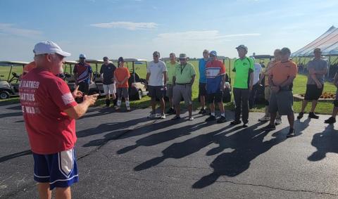 A group of people standing outdoors in a sunny area, listening to a speaker.