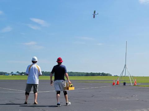 Two people watch a model plane flying in a clear blue sky over an open field.