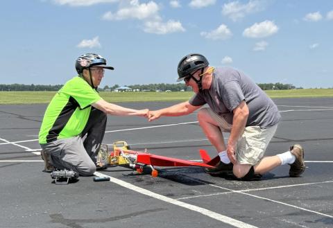 Two people wearing helmets fist bump on an airfield near a small red model plane.