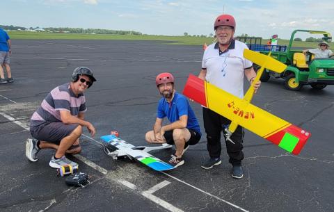 Three people with model airplanes on an airfield, smiling and wearing helmets.