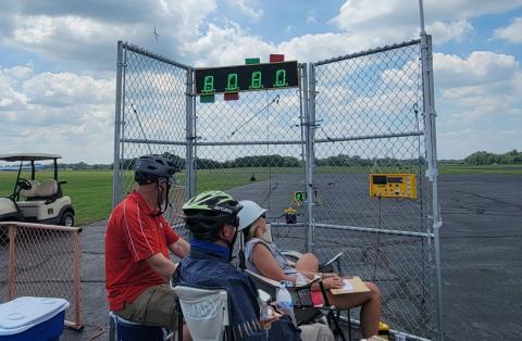 Three people in helmets watch a digital scoreboard at an outdoor event.