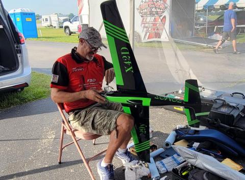 Man in red shirt working on a model aircraft with green accents outdoors.