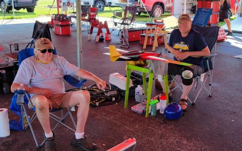 Two men relaxing in lawn chairs under a canopy, with model airplanes nearby.