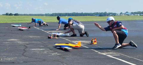 Model airplanes ready for takeoff, with people crouched nearby on an airstrip.
