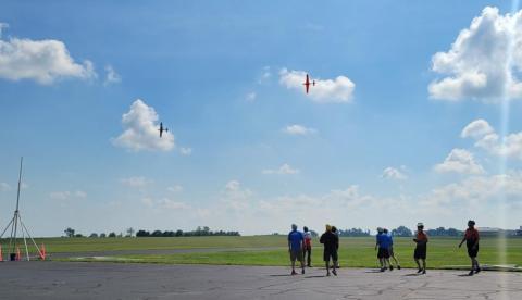 People watch flying model planes in a sunny open field.