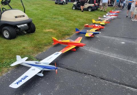 Model airplanes lined up on pavement, near grass, with people and a golf cart in the background.