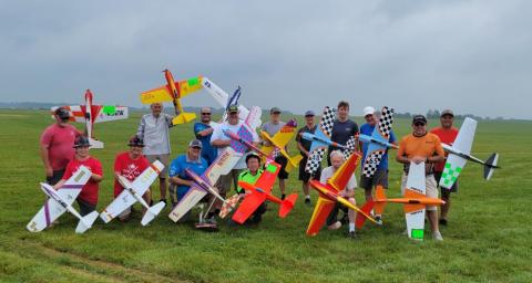 Group of people posing with colorful model airplanes in a grassy field.