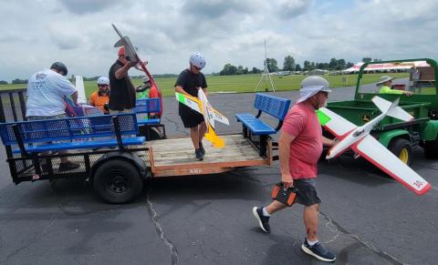 Men unloading remote-controlled airplanes from a blue and orange trailer on an overcast day.