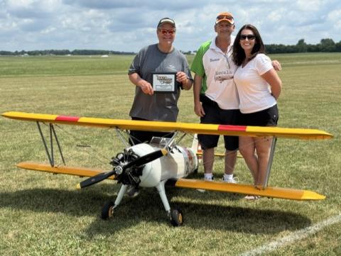 Three people smiling behind a yellow biplane model on a grassy field.