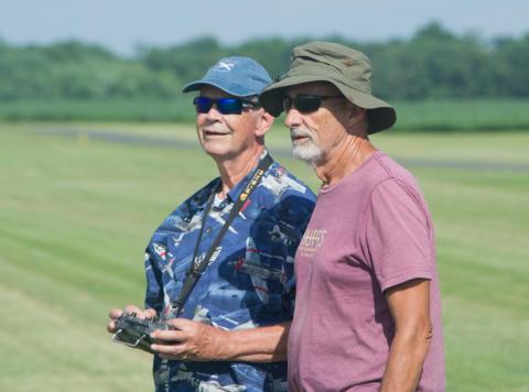 Two men wearing casual clothes and hats stand in a grassy field.