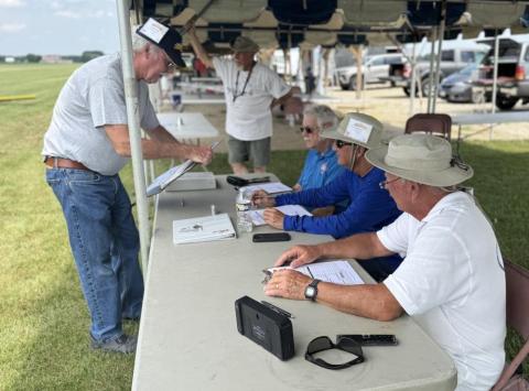 People sitting at a table outdoors, checking documents, beside parked cars.