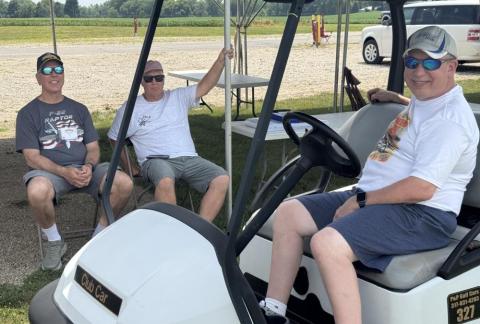 Three men sitting and relaxing by a golf cart under a tent.
