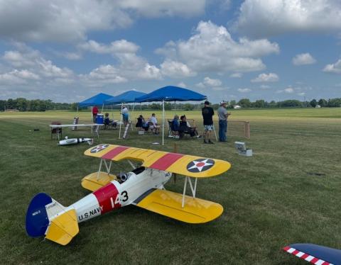 Model airplane with yellow wings on grassy field, people under blue tents.