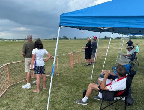 People watching model planes under blue tents on a grassy field.