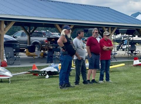 Four men standing in a row near model airplanes on grass.