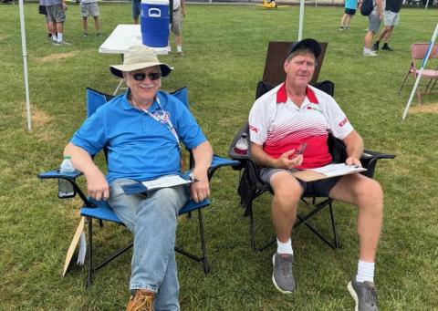 Two men sitting on lawn chairs under a canopy, casually dressed, in a grassy park.