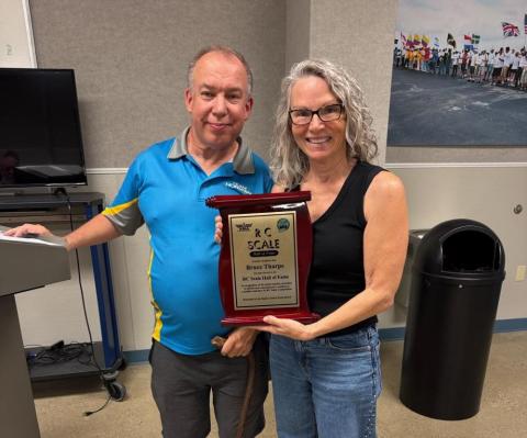 Two people smiling, holding a plaque in a room with a podium and wall decoration.