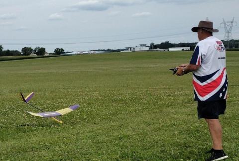 Man in shorts and hat flying remote-controlled glider in a grassy field.