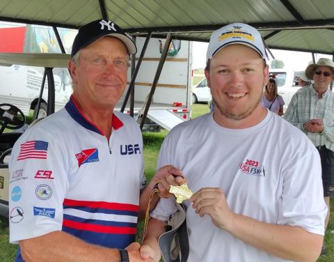 Two men smiling, one holding a gold medal, both wearing caps at an outdoor event.