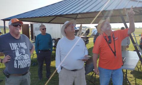 Four men stand outside under a metal canopy, one pointing upward, wearing casual clothes.