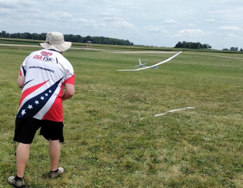 Man in hat flying a model glider over grassy field.