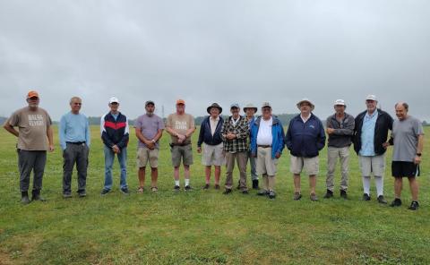 A group of twelve men standing on grass, dressed casually, under a cloudy sky.