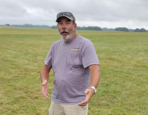 Man in a purple shirt standing in a grassy field, looking surprised.