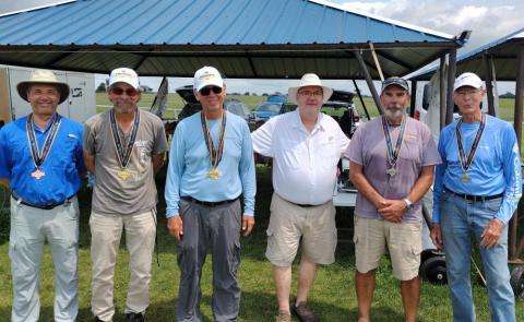 Six men standing under a metal-roofed pavilion, wearing hats and medals.