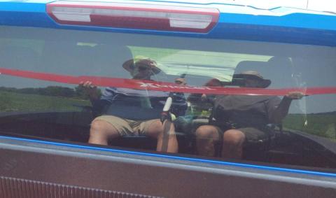 Two people with hats reflected in a car window, seated in front of a blurred landscape.