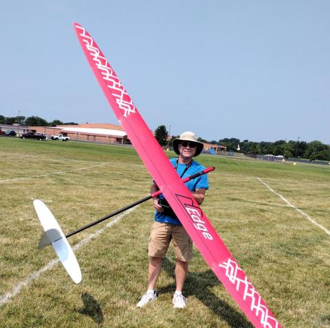 Man standing on grass holding a large pink model glider, wearing a hat and sunglasses.