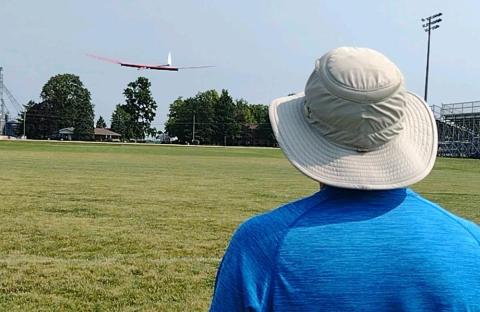 A person in a sun hat watches a glider flying over a grassy field.